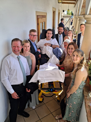 Eine Gruppe von mehreren Personen posiert zusammen am Eingang eines Gebäudes. Einige tragen elegante Kleidung, während ein Mann auf einer Trage sitzt, die von einer anderen Person geschoben wird. Die Szene wirkt festlich, möglicherweise bei einer Feier oder Hochzeit.