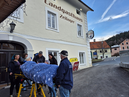 Eine Gruppe von Personen steht vor dem Landgasthaus Zeilinger. Sie helfen einer Person, die mit einer Decke bedeckt ist, auf eine Trage. Das Gebäude hat ein rustikales Aussehen, und die Umgebung ist ländlich.