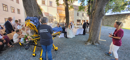 Hochzeitsfeier im Freien mit einem Rettungswagen und Gästen im Hintergrund.