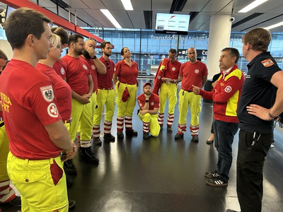 Eine Gruppe von Rettungskräften in rot-gelben Uniformen versammelt sich in einem modernen Raum. Ein Teamleiter spricht. Einige Teammitglieder hören aufmerksam zu, während andere eine entspannte Haltung einnehmen. Im Hintergrund sind große Fenster mit Stadtansicht sichtbar.