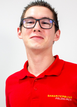 Junger Mann in rotem Polohemd lächelt, trägt Brille und hat kurzes Haar.