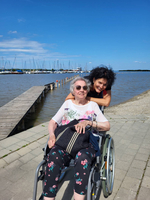 Eine ältere Frau im Rollstuhl mit einer Frau, die sie umarmt, am Wasser.