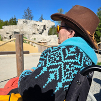 Eine Person mit einem Hut und einer bunten Jacke sitzt auf einem Rollstuhl in der Natur. Im Hintergrund ist die steinige Struktur der Westernstadt zu sehen.