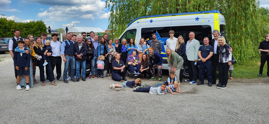 Eine große Gruppe von Menschen steht zusammen auf einem Parkplatz neben einem Lieferwagen mit buntem Design. Es sind Erwachsene und Kinder unterschiedlichen Alters. Einige Kinder liegen auf dem Boden, während andere stehen oder sitzen. Im Hintergrund sind Bäume und ein blauer Himmel mit Wolken zu sehen.