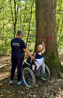 Eine Person im Rollstuhl berührt einen Baum, während eine Begleitperson hilft.