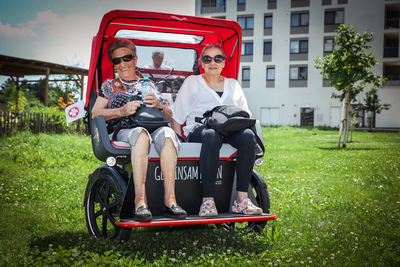 zwei ältere Damen aus Senioren WG sitzen in Samariterbund Wien Rikscha, beide tragen eine Sonnenbrille und lächeln in die Kamera, Rikscha steht auf Grünfläche, dahinter Gebäude